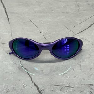 May include: Purple sunglasses with blue mirrored lenses. The frames are a solid, matte purple. The lenses reflect the surrounding environment. The sunglasses have a sleek, modern design.