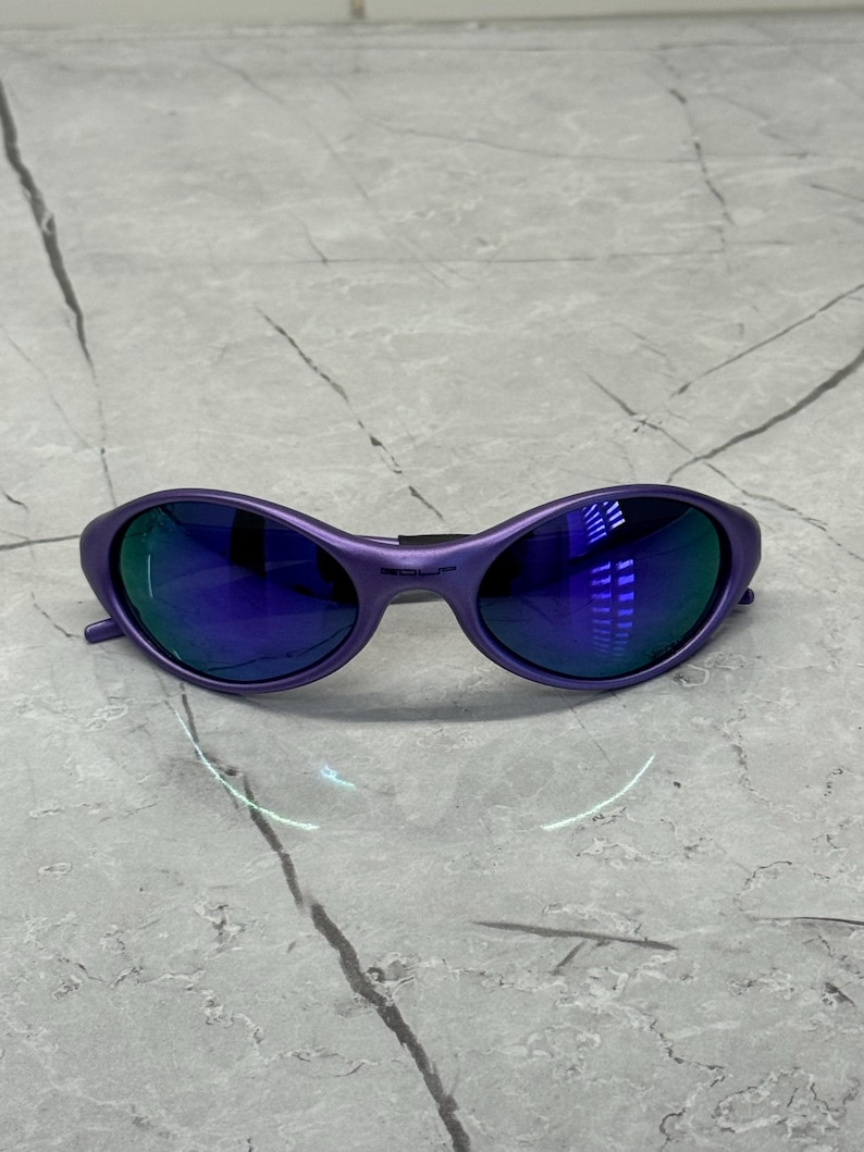 May include: Purple sunglasses with blue mirrored lenses. The frames are a solid, matte purple. The lenses reflect the surrounding environment. The sunglasses have a sleek, modern design.