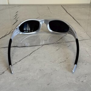 May include: Silver-framed sunglasses with black lenses. The sunglasses have a sleek, modern design with a curved frame and black temples. The lenses are dark, providing shade and protection from the sun.