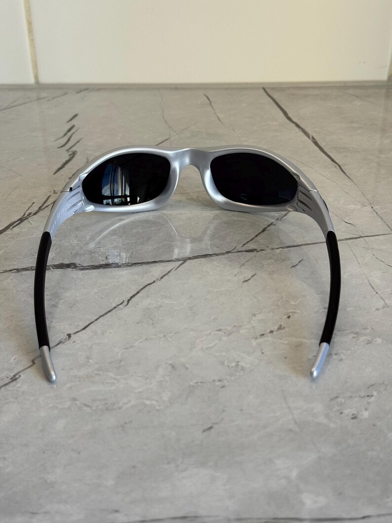 May include: Silver-framed sunglasses with black lenses. The sunglasses have a sleek, modern design with a curved frame and black temples. The lenses are dark, providing shade and protection from the sun.