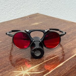 May include: Black sunglasses with round, red-tinted lenses and a central, cylindrical attachment. The glasses have a sleek, modern design, and are set against a wooden surface.