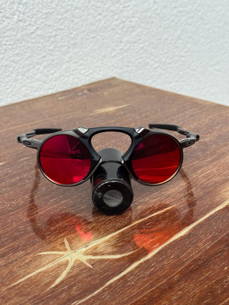 May include: Black sunglasses with round, red-tinted lenses and a central, cylindrical attachment. The glasses have a sleek, modern design, and are set against a wooden surface.