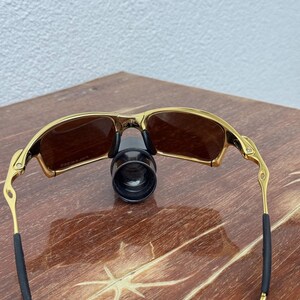 May include: Gold-framed sunglasses with brown lenses. The sunglasses have a sleek, modern design with a gold finish on the frame and temples. The arms have black rubber grips for comfort. The lenses are a warm brown color.