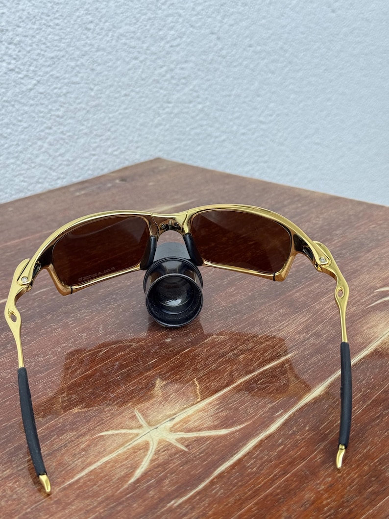 May include: Gold-framed sunglasses with brown lenses. The sunglasses have a sleek, modern design with a gold finish on the frame and temples. The arms have black rubber grips for comfort. The lenses are a warm brown color.