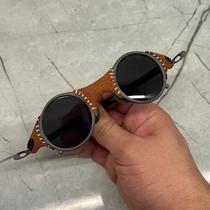 May include: Round sunglasses with dark lenses and a brown leather bridge. The frame is a dark gray color, and the leather is stitched with white thread. The sunglasses have a unique, vintage style.
