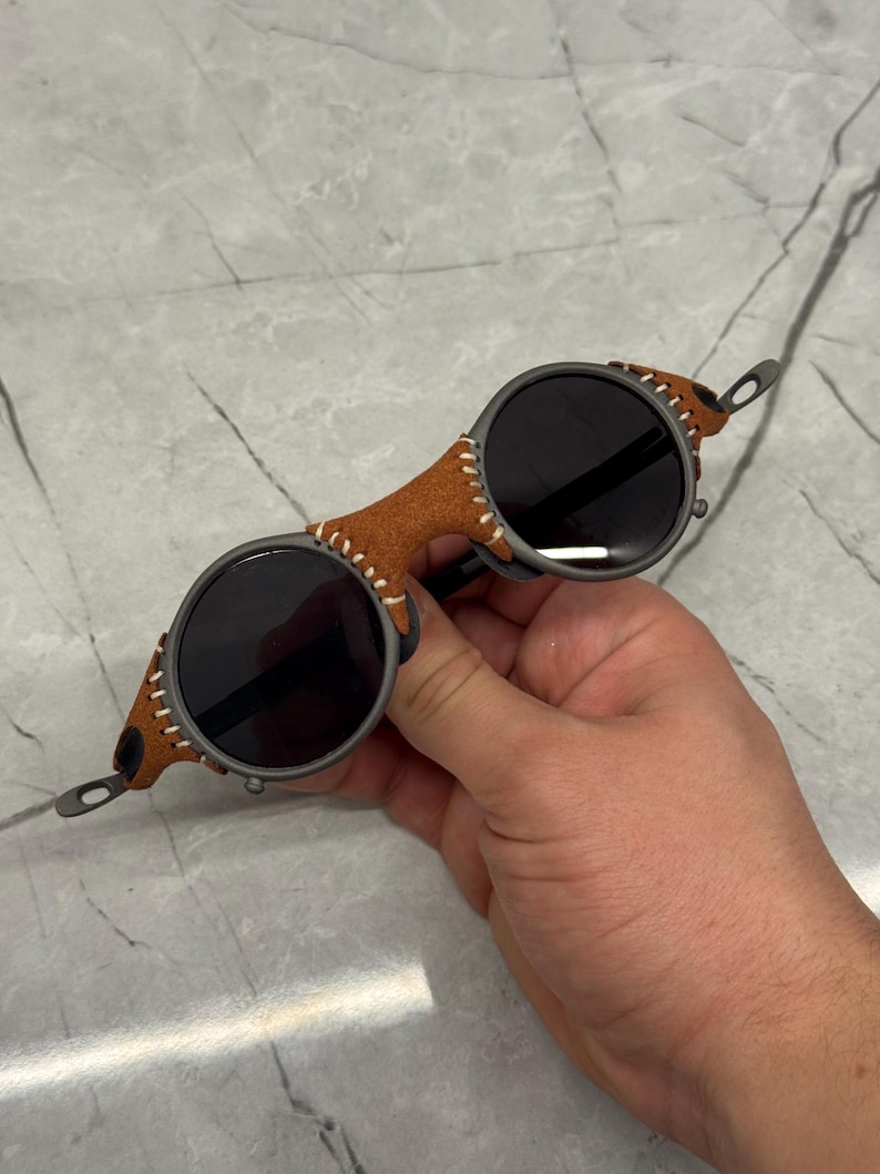 May include: Round sunglasses with dark lenses and a brown leather bridge. The frame is a dark gray color, and the leather is stitched with white thread. The sunglasses have a unique, vintage style.