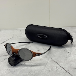 May include: A pair of round-lens sunglasses with a brown leather accent and a black case. The sunglasses have a dark gray frame and lenses. The case is black and has the brand name "Oakley" on it.