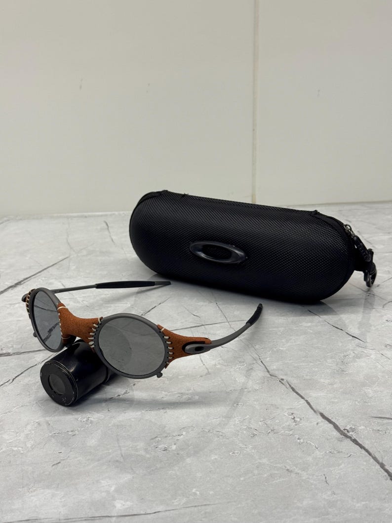 May include: A pair of round-lens sunglasses with a brown leather accent and a black case. The sunglasses have a dark gray frame and lenses. The case is black and has the brand name "Oakley" on it.