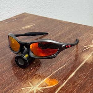 Op de afbeelding: Een zwarte zonnebril met oranje-rode glazen ligt op een houten oppervlak. De zonnebril heeft een gebogen ontwerp en een klein rood logo op de poot. De glazen reflecteren licht, waardoor een levendig effect ontstaat. Het houten oppervlak heeft een natuurlijke nerf.