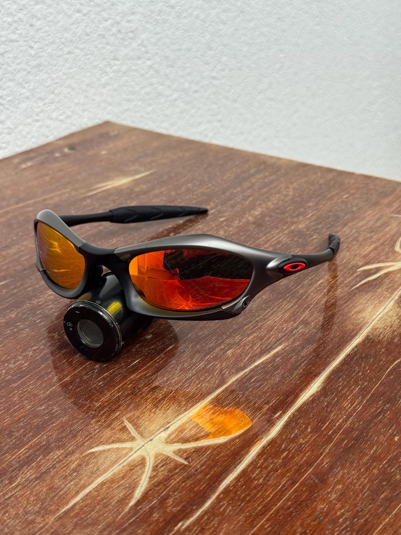 Op de afbeelding: Een zwarte zonnebril met oranje-rode glazen ligt op een houten oppervlak. De zonnebril heeft een gebogen ontwerp en een klein rood logo op de poot. De glazen reflecteren licht, waardoor een levendig effect ontstaat. Het houten oppervlak heeft een natuurlijke nerf.