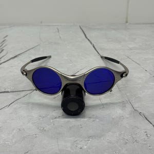 May include: Silver-framed sunglasses with round blue lenses. The sunglasses have a unique design with a nose bridge and black earpieces. The lenses are a vibrant blue color.