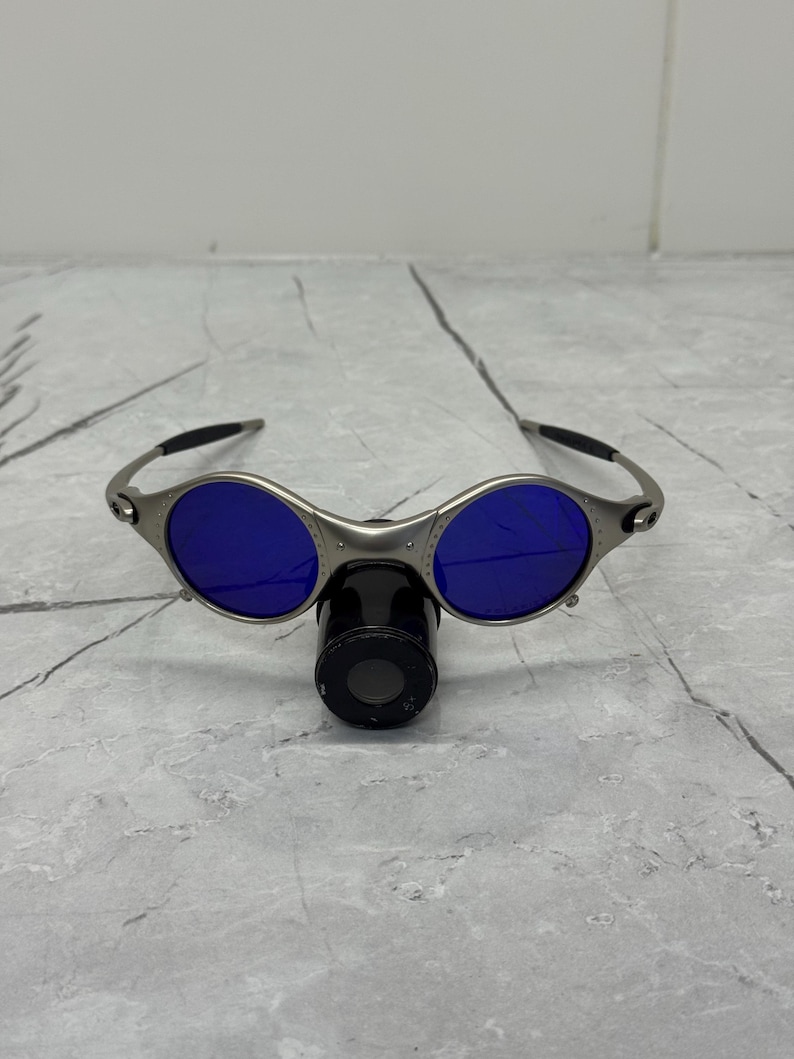 May include: Silver-framed sunglasses with round blue lenses. The sunglasses have a unique design with a nose bridge and black earpieces. The lenses are a vibrant blue color.
