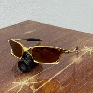 May include: Gold-framed sunglasses with brown lenses. The sunglasses have a rectangular shape and a sleek, modern design. The frame is a shiny gold color, and the lenses are a warm brown. The sunglasses are resting on a wooden surface.
