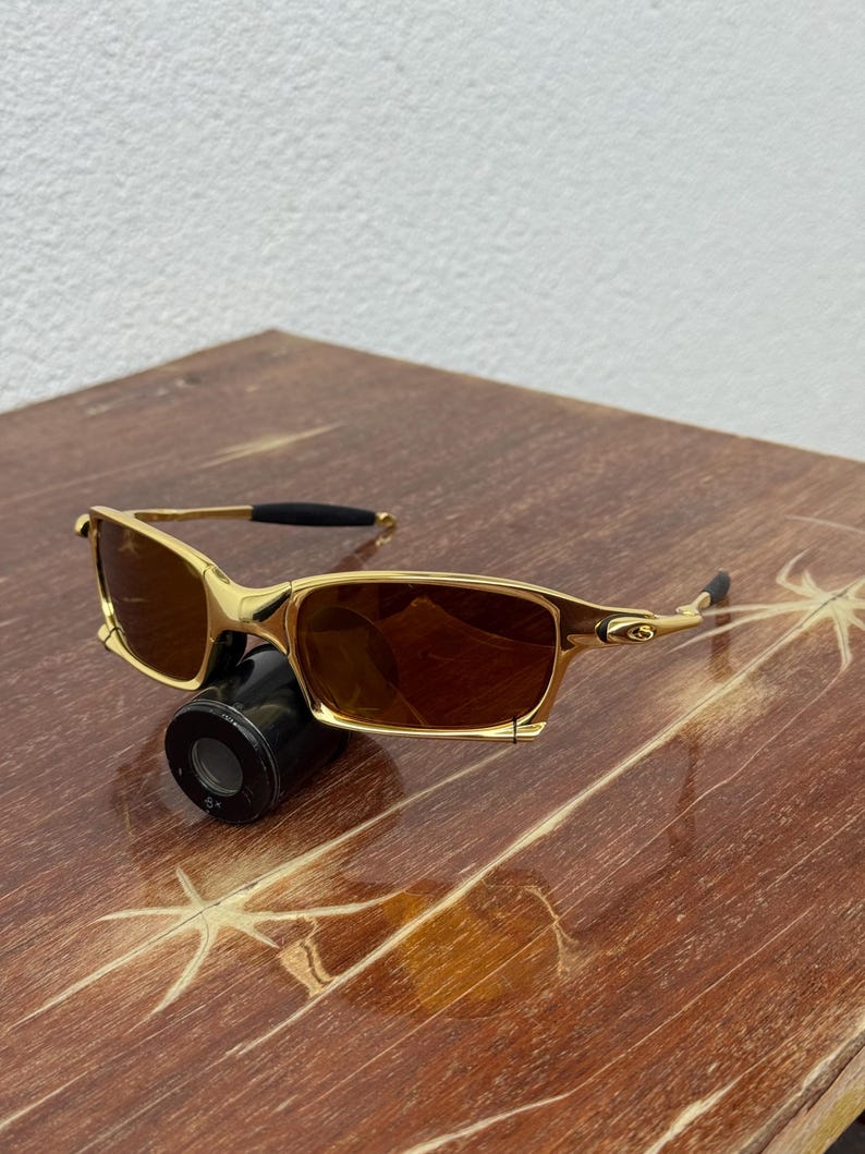 May include: Gold-framed sunglasses with brown lenses. The sunglasses have a rectangular shape and a sleek, modern design. The frame is a shiny gold color, and the lenses are a warm brown. The sunglasses are resting on a wooden surface.