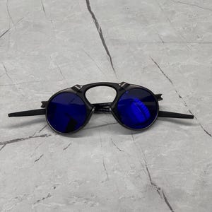 以下が含まれることがあります： 丸い青色のレンズが付いた黒いサングラス。 サングラスは、レンズから伸びるブリッジとテンプルを備えたユニークなデザインです。 レンズは鮮やかな青色です。