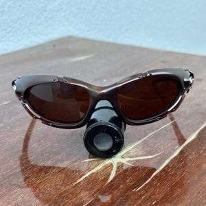 Puede incluir: Gafas de sol marrones con montura curvada y lentes oscuras. Las gafas de sol est&aacute;n sobre una superficie de madera. Una peque&ntilde;a lupa negra est&aacute; colocada debajo.