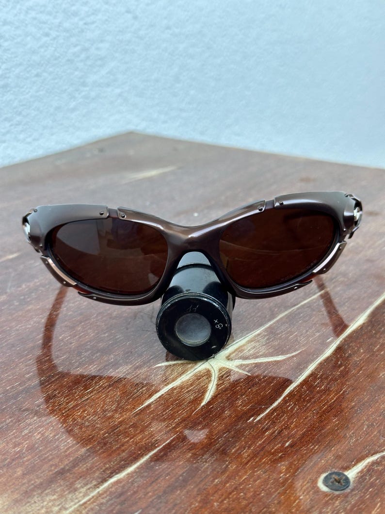 Puede incluir: Gafas de sol marrones con montura curvada y lentes oscuras. Las gafas de sol est&aacute;n sobre una superficie de madera. Una peque&ntilde;a lupa negra est&aacute; colocada debajo.