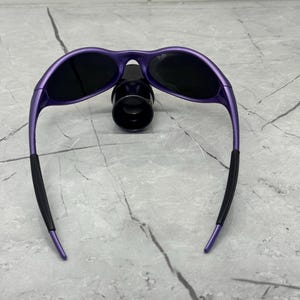 May include: Purple sunglasses with black lenses and black rubber ear pieces. The sunglasses have a sleek, curved design and are resting on a black object. The frame is a vibrant purple color.