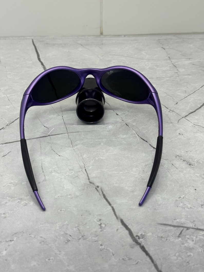 May include: Purple sunglasses with black lenses and black rubber ear pieces. The sunglasses have a sleek, curved design and are resting on a black object. The frame is a vibrant purple color.