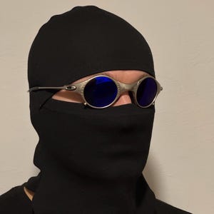 May include: Silver-framed sunglasses with round, blue lenses. The sunglasses are worn with a black balaclava and a black garment covering the lower face and neck. The background is a plain, light-colored wall.