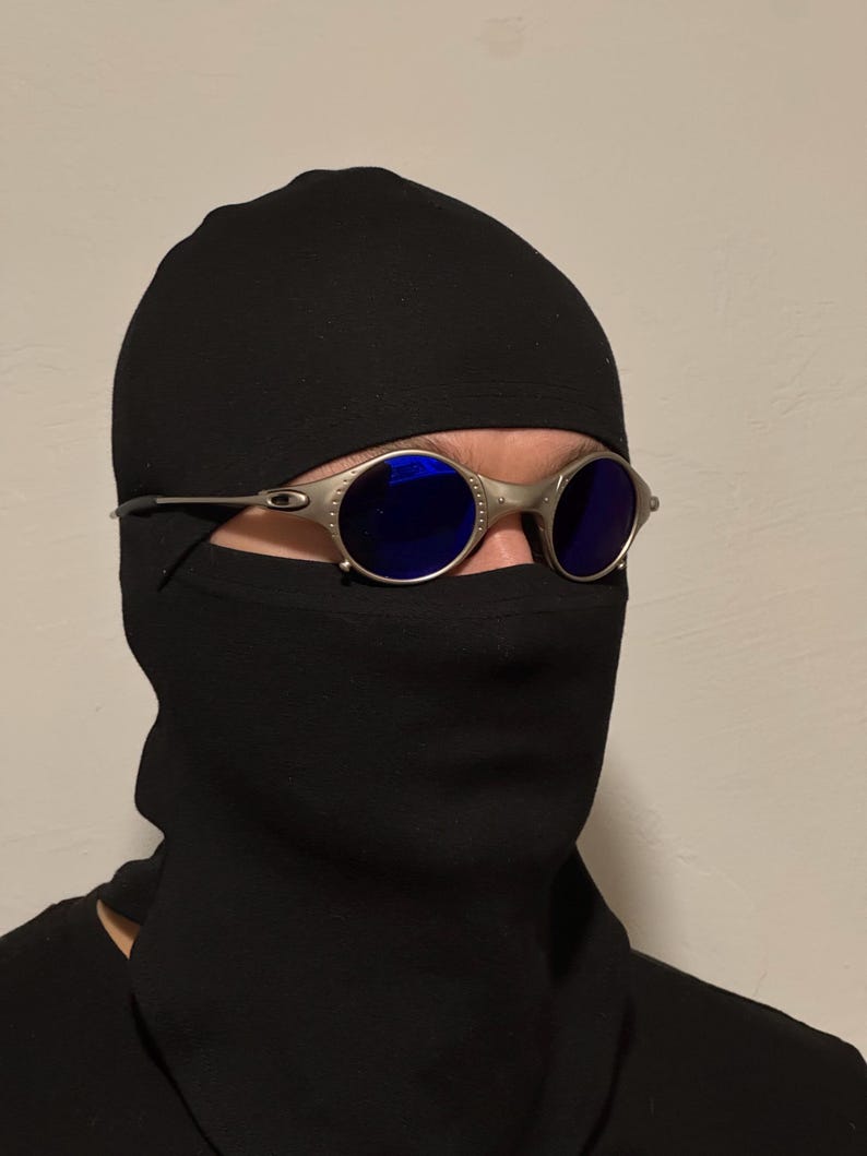 May include: Silver-framed sunglasses with round, blue lenses. The sunglasses are worn with a black balaclava and a black garment covering the lower face and neck. The background is a plain, light-colored wall.
