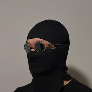 May include: A black balaclava with a face covering and a pair of round, dark-tinted sunglasses. The sunglasses have a thin, silver frame and a brown bridge. The balaclava covers the head and neck.