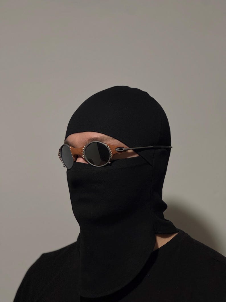 May include: A black balaclava with a face covering and a pair of round, dark-tinted sunglasses. The sunglasses have a thin, silver frame and a brown bridge. The balaclava covers the head and neck.