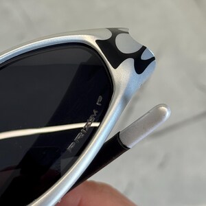 May include: Silver sunglasses with black lenses. The frame features a black and white flame design on the temples. The text "PRIZM P" is visible on the lens. The sunglasses have a sleek, modern design.