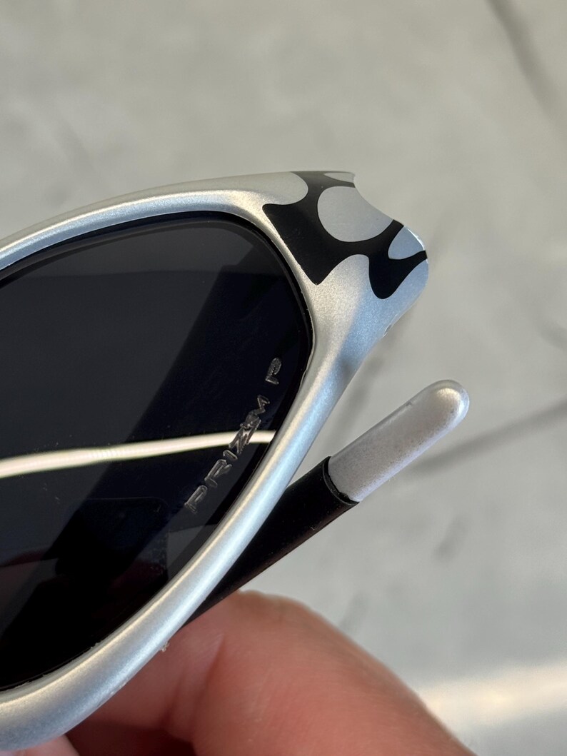 May include: Silver sunglasses with black lenses. The frame features a black and white flame design on the temples. The text "PRIZM P" is visible on the lens. The sunglasses have a sleek, modern design.