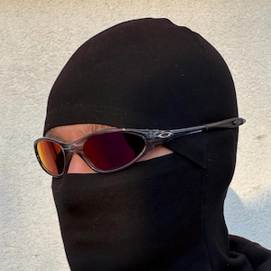 K&ouml;nnte beinhalten: Eine Sonnenbrille mit transparentem Rahmen und rot get&ouml;nten Gl&auml;sern wird &uuml;ber einer schwarzen Sturmhaube getragen. Die Sonnenbrille hat ein sportliches Design, und die Sturmhaube bedeckt Kopf und Hals, wobei nur die Augen sichtbar sind.