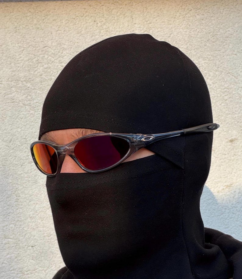 K&ouml;nnte beinhalten: Eine Sonnenbrille mit transparentem Rahmen und rot get&ouml;nten Gl&auml;sern wird &uuml;ber einer schwarzen Sturmhaube getragen. Die Sonnenbrille hat ein sportliches Design, und die Sturmhaube bedeckt Kopf und Hals, wobei nur die Augen sichtbar sind.