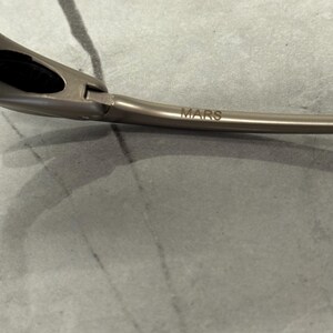 May include: Close-up of a pair of sunglasses with a metallic frame. The word "MARS" is printed on the arm of the glasses. The glasses have a sleek, modern design.