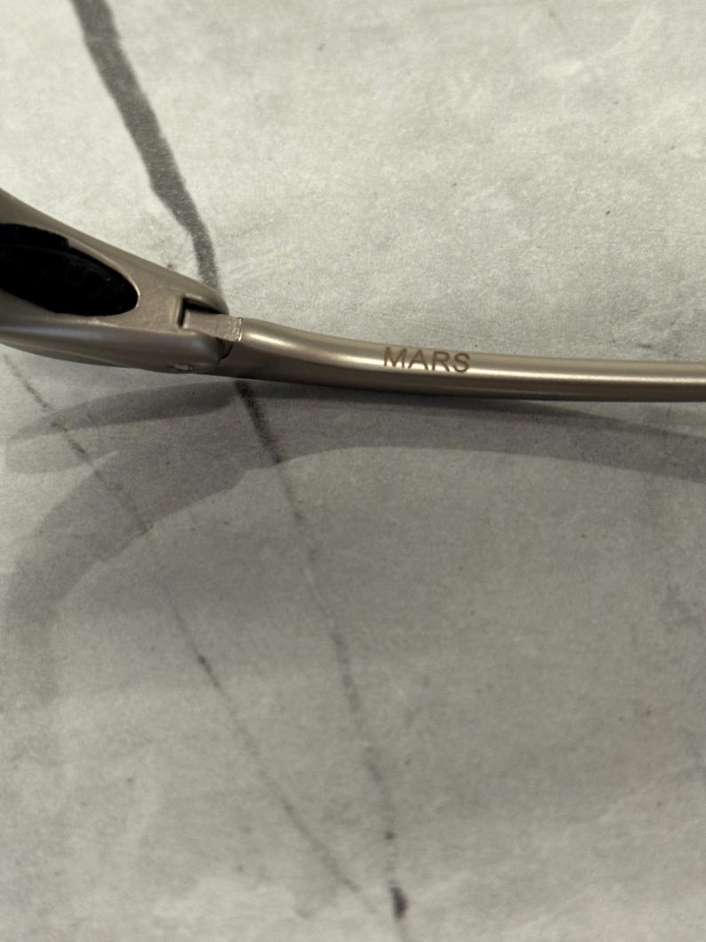 May include: Close-up of a pair of sunglasses with a metallic frame. The word "MARS" is printed on the arm of the glasses. The glasses have a sleek, modern design.