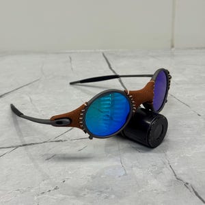 以下が含まれることがあります： 青いミラーレンズと茶色の革のアクセントが付いた楕円形のサングラス。フレームはダークグレーで、曲線的なデザインです。サングラスは黒い円筒形の物体の上に置かれています。
