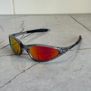 K&ouml;nnte beinhalten: Graue Sonnenbrille mit roten und gelben Gl&auml;sern. Das Wrap-around-Design hat einen schwarzen B&uuml;gel und einen grauen Rahmen. Die Gl&auml;ser haben einen Farbverlauf, der von Gelb nach Rot verl&auml;uft.