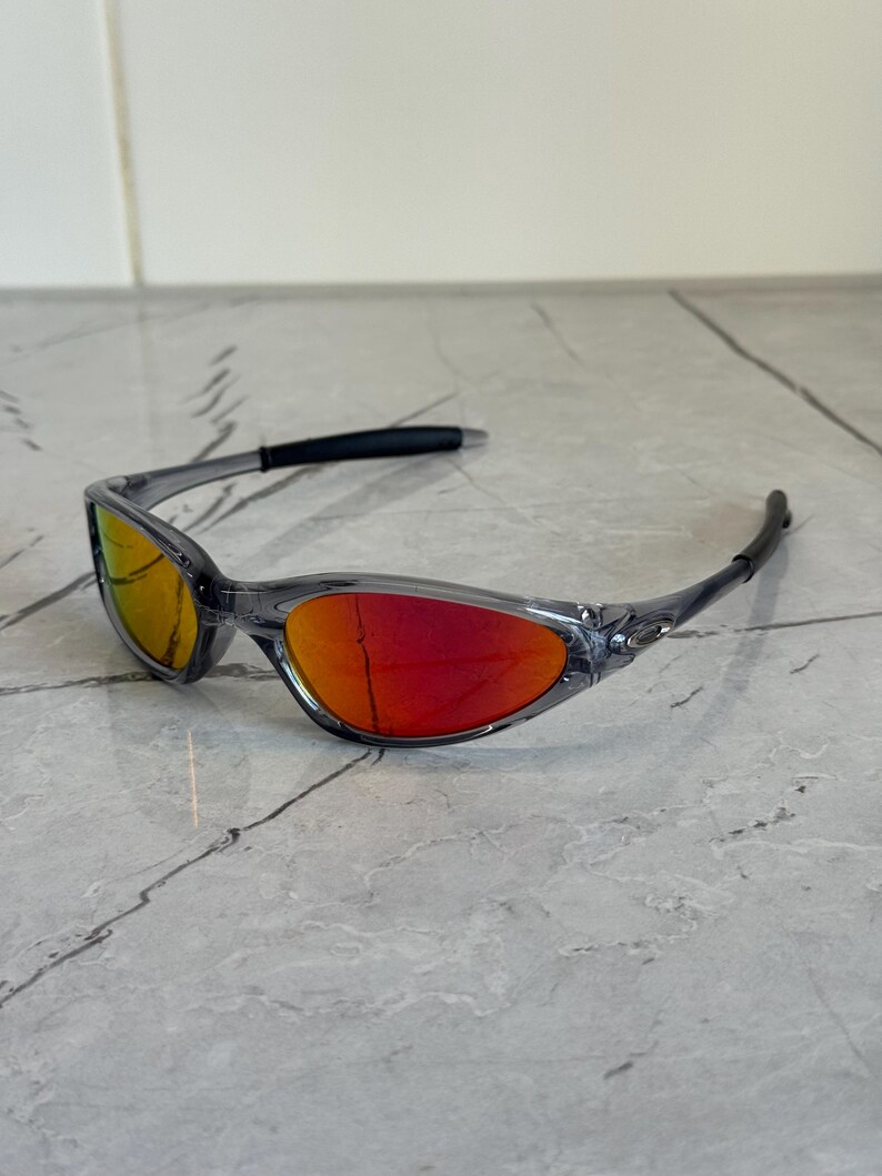 K&ouml;nnte beinhalten: Graue Sonnenbrille mit roten und gelben Gl&auml;sern. Das Wrap-around-Design hat einen schwarzen B&uuml;gel und einen grauen Rahmen. Die Gl&auml;ser haben einen Farbverlauf, der von Gelb nach Rot verl&auml;uft.