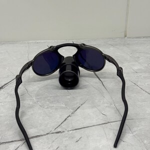 May include: Black sunglasses with dark blue lenses and a unique design. The sunglasses have a central cylindrical component and curved arms. The frame appears to be made of metal.