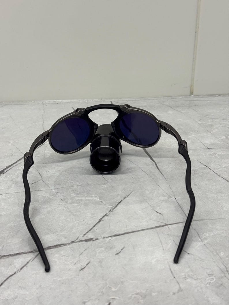 May include: Black sunglasses with dark blue lenses and a unique design. The sunglasses have a central cylindrical component and curved arms. The frame appears to be made of metal.