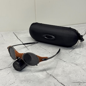 May include: Black sunglasses with brown leather accents and a black case. The sunglasses have a round frame and dark lenses. The case is oval-shaped with a zipper closure and the Oakley logo.
