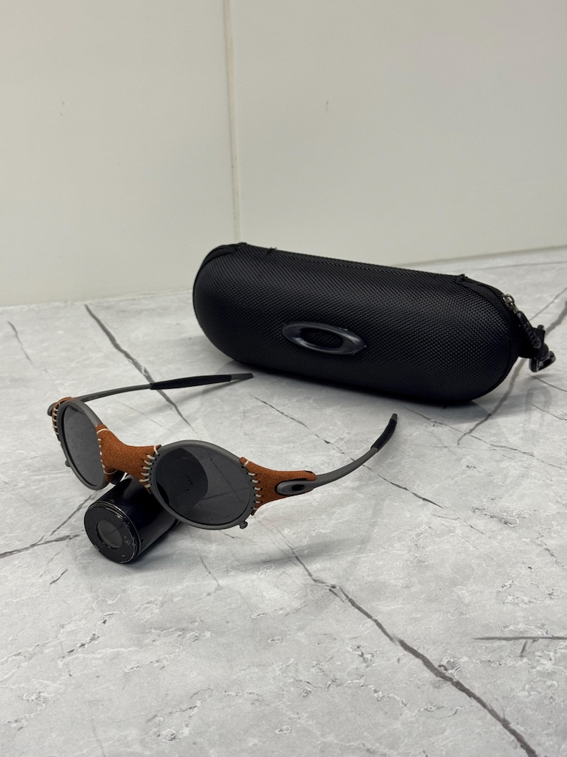 May include: Black sunglasses with brown leather accents and a black case. The sunglasses have a round frame and dark lenses. The case is oval-shaped with a zipper closure and the Oakley logo.