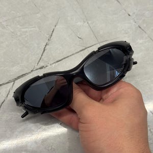 May include: Black sunglasses with a unique, textured frame design. The lenses are dark, and the frame has a series of small, raised details. The sunglasses are held in a hand, showcasing their shape and style.