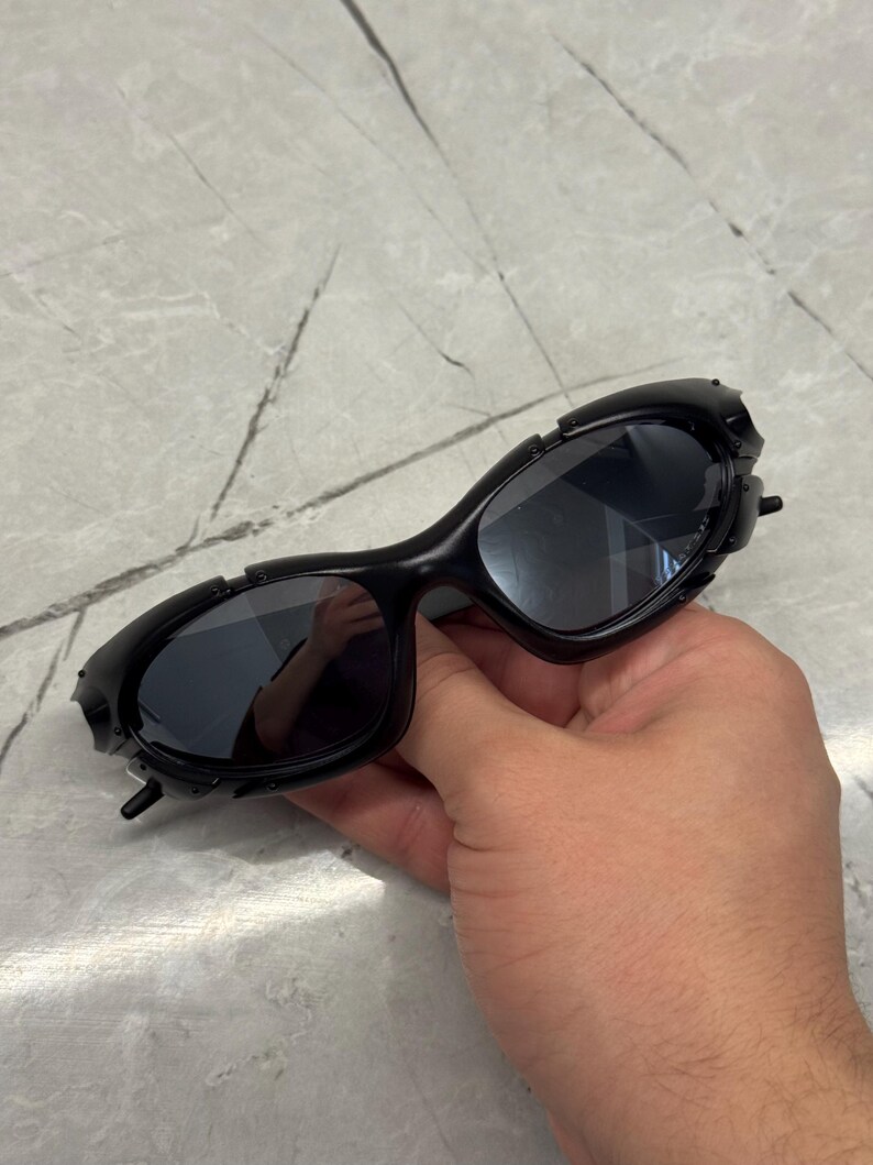 May include: Black sunglasses with a unique, textured frame design. The lenses are dark, and the frame has a series of small, raised details. The sunglasses are held in a hand, showcasing their shape and style.