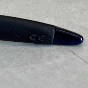 K&ouml;nnte beinhalten: Nahaufnahme des Endes eines schwarzen Brillenb&uuml;gels mit einer dunkelblauen Spitze. Der B&uuml;gel hat einen gummierten Abschnitt in der N&auml;he des Scharniers, und die Buchstaben "CE" sind sichtbar.
