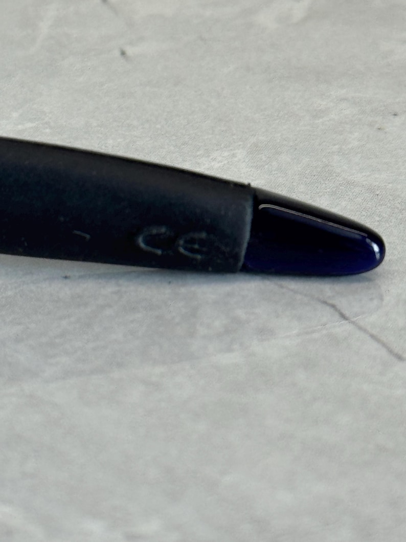K&ouml;nnte beinhalten: Nahaufnahme des Endes eines schwarzen Brillenb&uuml;gels mit einer dunkelblauen Spitze. Der B&uuml;gel hat einen gummierten Abschnitt in der N&auml;he des Scharniers, und die Buchstaben "CE" sind sichtbar.
