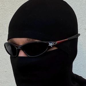 K&ouml;nnte beinhalten: Schwarze Sonnenbrille mit Sternlogo an den B&uuml;geln, getragen mit einer schwarzen Sturmhaube. Die Sonnenbrille hat ein schlankes, sportliches Design mit dunklen Gl&auml;sern und einem roten Akzent an den B&uuml;geln. Die Sturmhaube bedeckt Kopf und Hals.