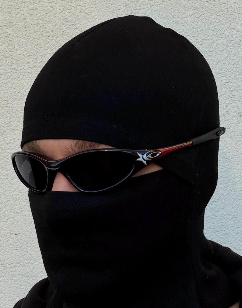 K&ouml;nnte beinhalten: Schwarze Sonnenbrille mit Sternlogo an den B&uuml;geln, getragen mit einer schwarzen Sturmhaube. Die Sonnenbrille hat ein schlankes, sportliches Design mit dunklen Gl&auml;sern und einem roten Akzent an den B&uuml;geln. Die Sturmhaube bedeckt Kopf und Hals.