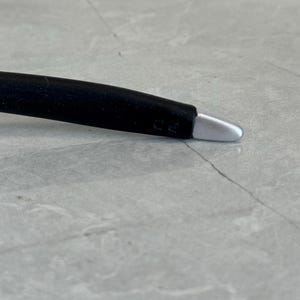 May include: A black stylus with a silver tip. The stylus is designed for use with touch screen devices. The stylus is lying on a grey surface.