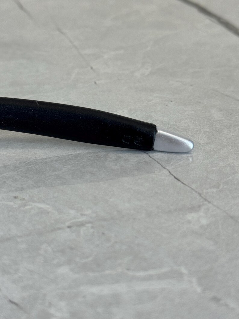 May include: A black stylus with a silver tip. The stylus is designed for use with touch screen devices. The stylus is lying on a grey surface.