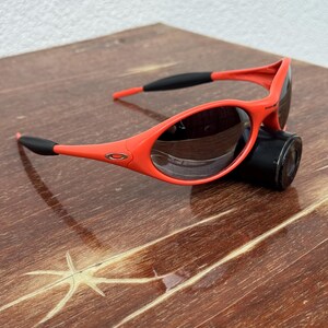 Pu&ograve; includere: Occhiali da sole arancioni vivaci con dettagli neri sulle aste. Questi occhiali sportivi presentano un design elegante e curvo e lenti scure. Gli occhiali da sole sono appoggiati su una superficie scura.