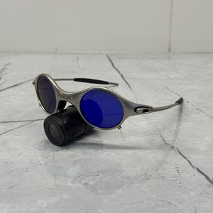 May include: Silver-framed sunglasses with blue lenses rest on a black cylindrical case. The sunglasses have a sleek, modern design with a curved frame and dark temples. The lenses are a vibrant blue color.